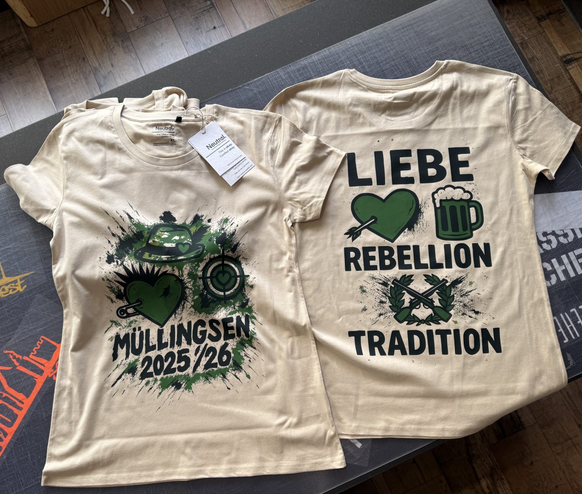 Zwei frisch bedruckte T-Shirts mit gr&uuml;n-schwarzen Motiven f&uuml;r M&uuml;llingsen 2025/26 auf dem Arbeitstisch der Hellweg Druckerei